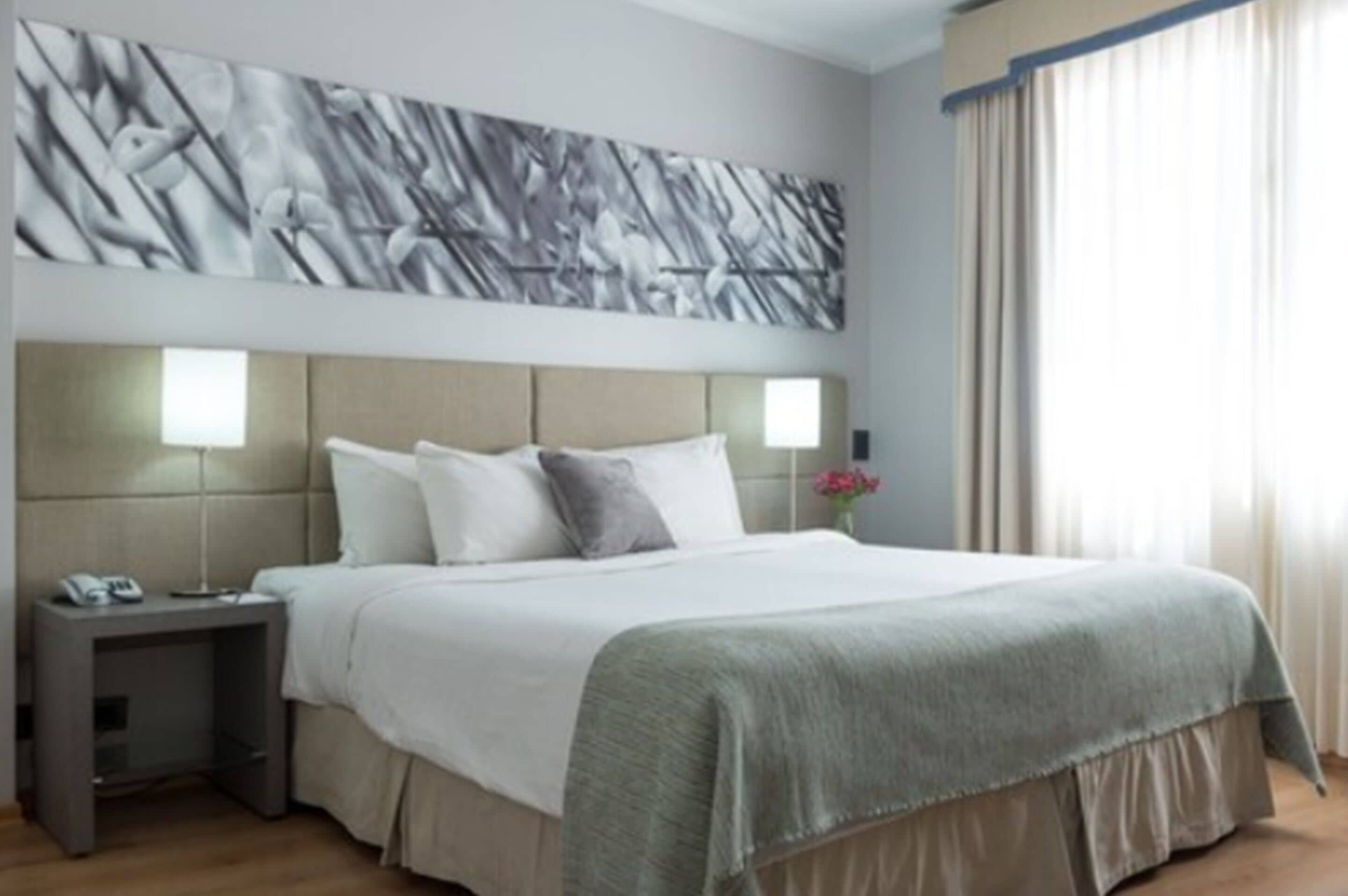 Moderni hotellihuone, jossa on suuri sänky, sängynpääty, jossa on integroitu valaistus, yöpöytä, jossa on puhelin, ja seinälle asennettu abstrakti taideteos.
