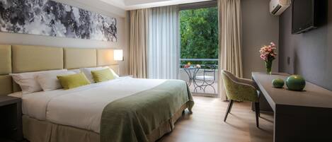 Habitación de hotel con cama, escritorio, silla, televisor y balcón con vistas a follaje.