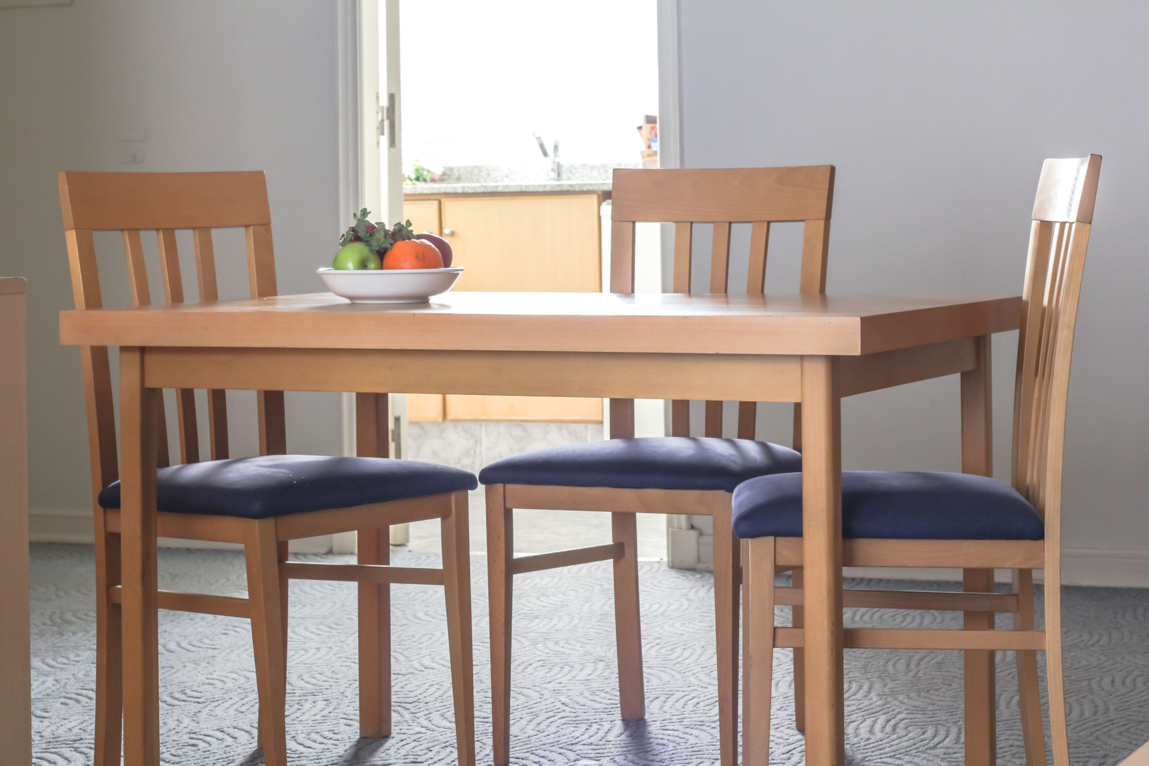 Una mesa de comedor con cuatro sillas de madera, un cuenco de fruta y una ventana al fondo.