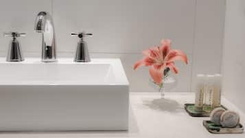 Un baño con un lavabo blanco, tres grifos cromados, un florero de vidrio con un lirio rosado y tres artículos de El Portal sobre una bandeja.