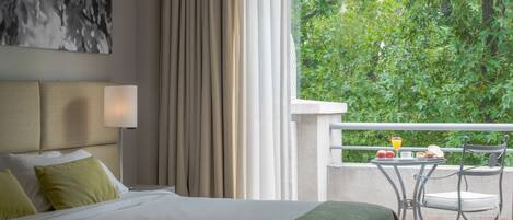 A hotel room with a bed, a bedside lamp, a view of greenery through the window, and a balcony with a table and chair.