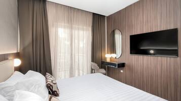 Habitación de hotel con cama, televisor de pantalla plana en la pared, mesita de noche con lámpara y ventana con cortinas traslúcidas.