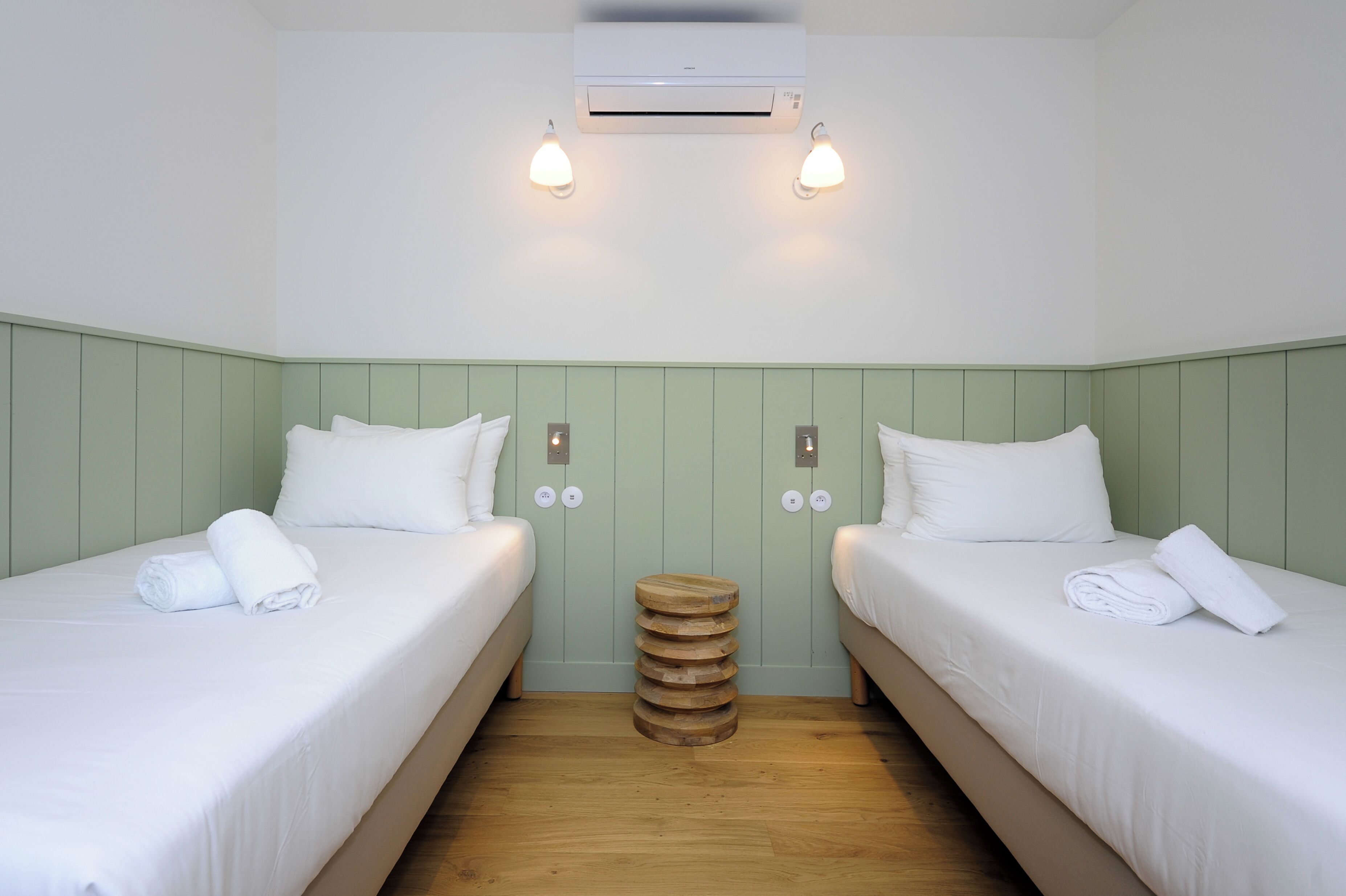 Habitación de hotel con dos camas, un taburete de madera en el centro y un equipo de aire acondicionado.