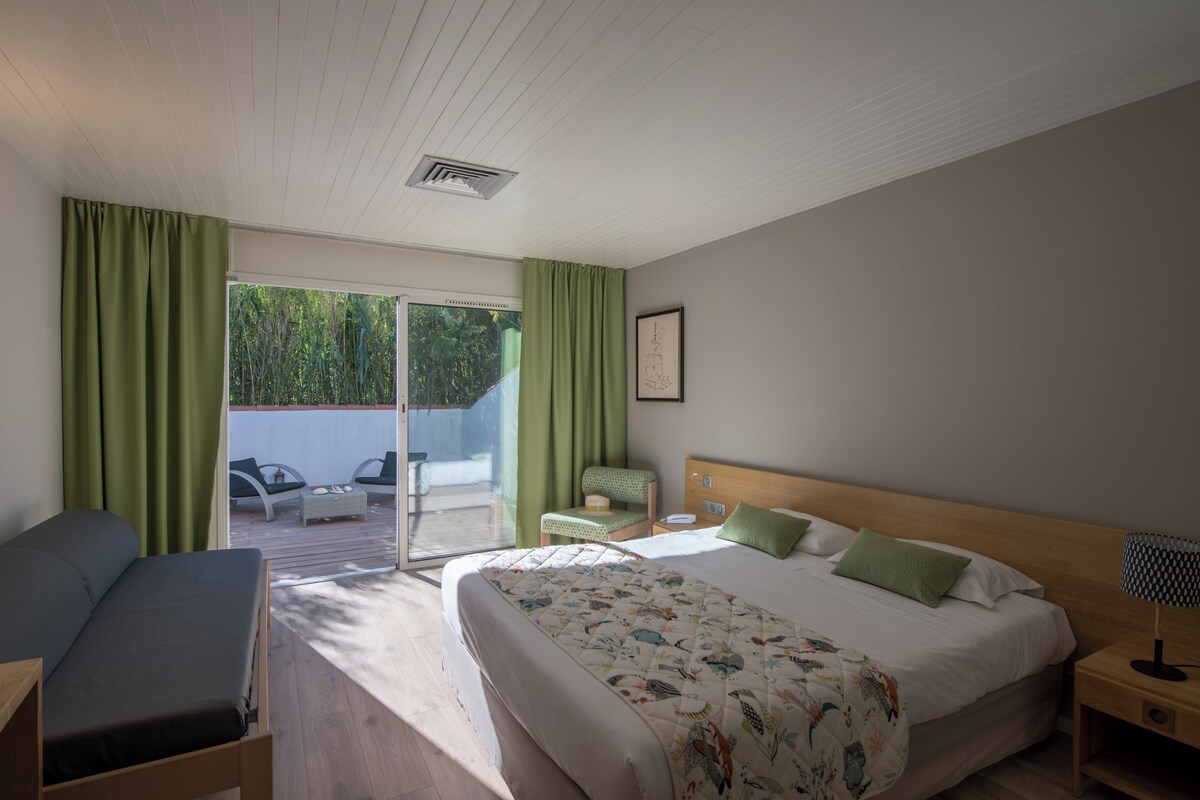 A bedroom with a bed, a chair, a small table, a nightstand, and a view of the outdoors.