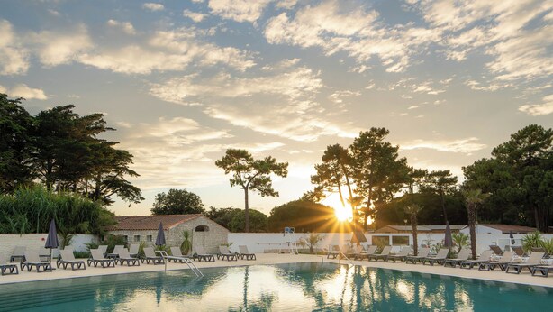 A swimming pool with lounge chairs and umbrellas, surrounded by trees and a building with a tiled roof. - Hotel Les Grenettes (Sainte-Marie-de-Re)