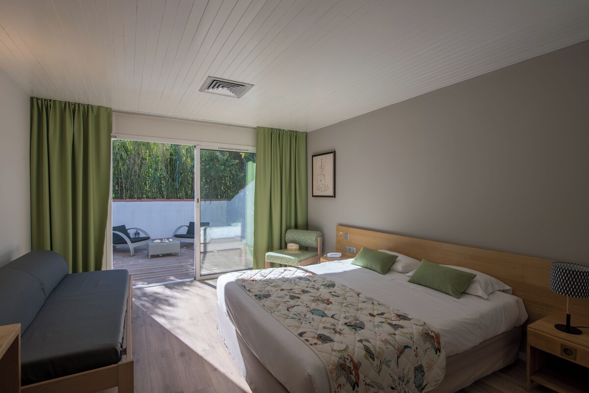 A bedroom with a bed, a chair, a small table, a nightstand, and a view of the outdoors.
