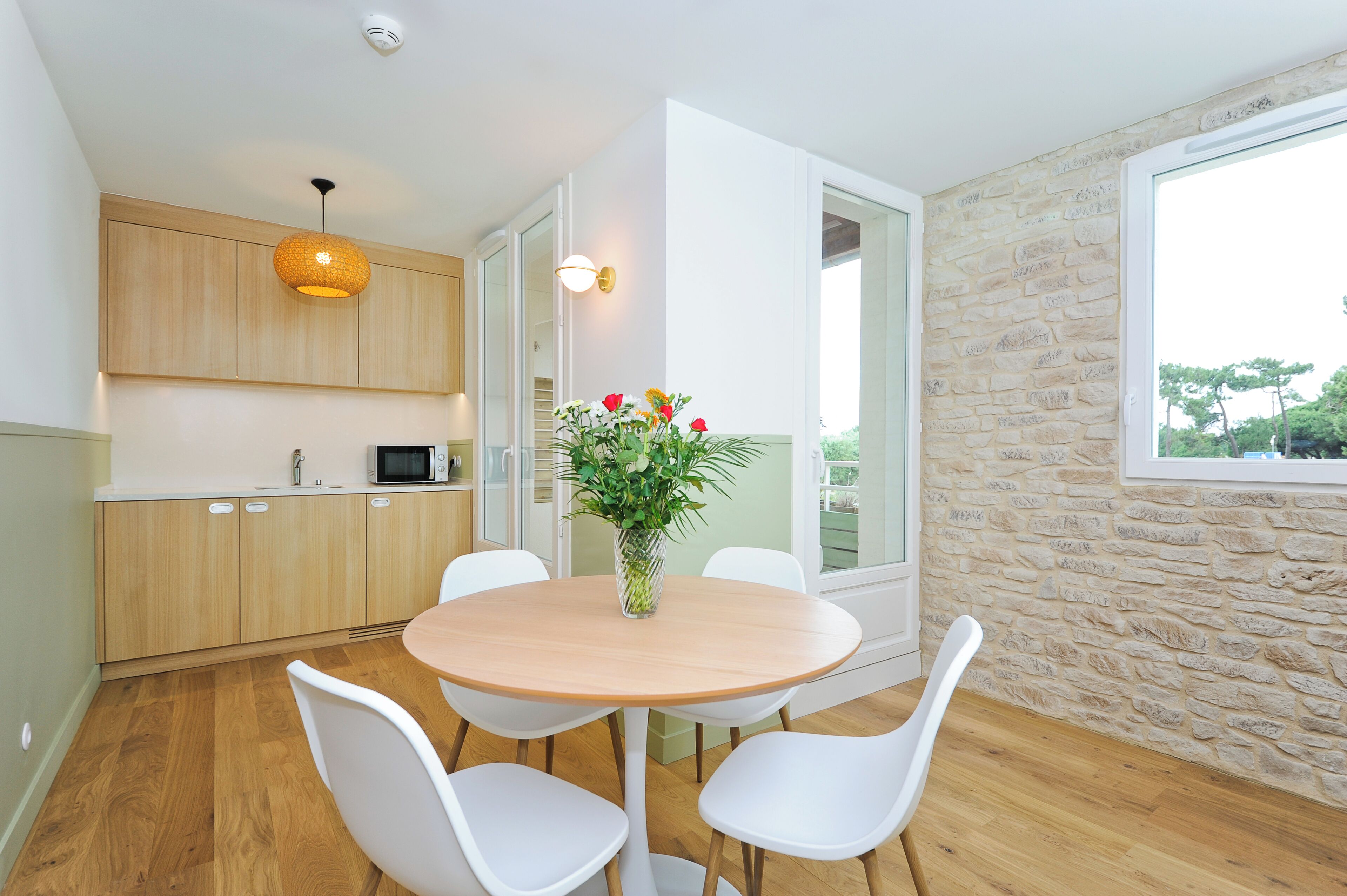 A modern kitchen with wooden cabinets, a round wooden table, white chairs, a potted plant, and a stone wall with a window.