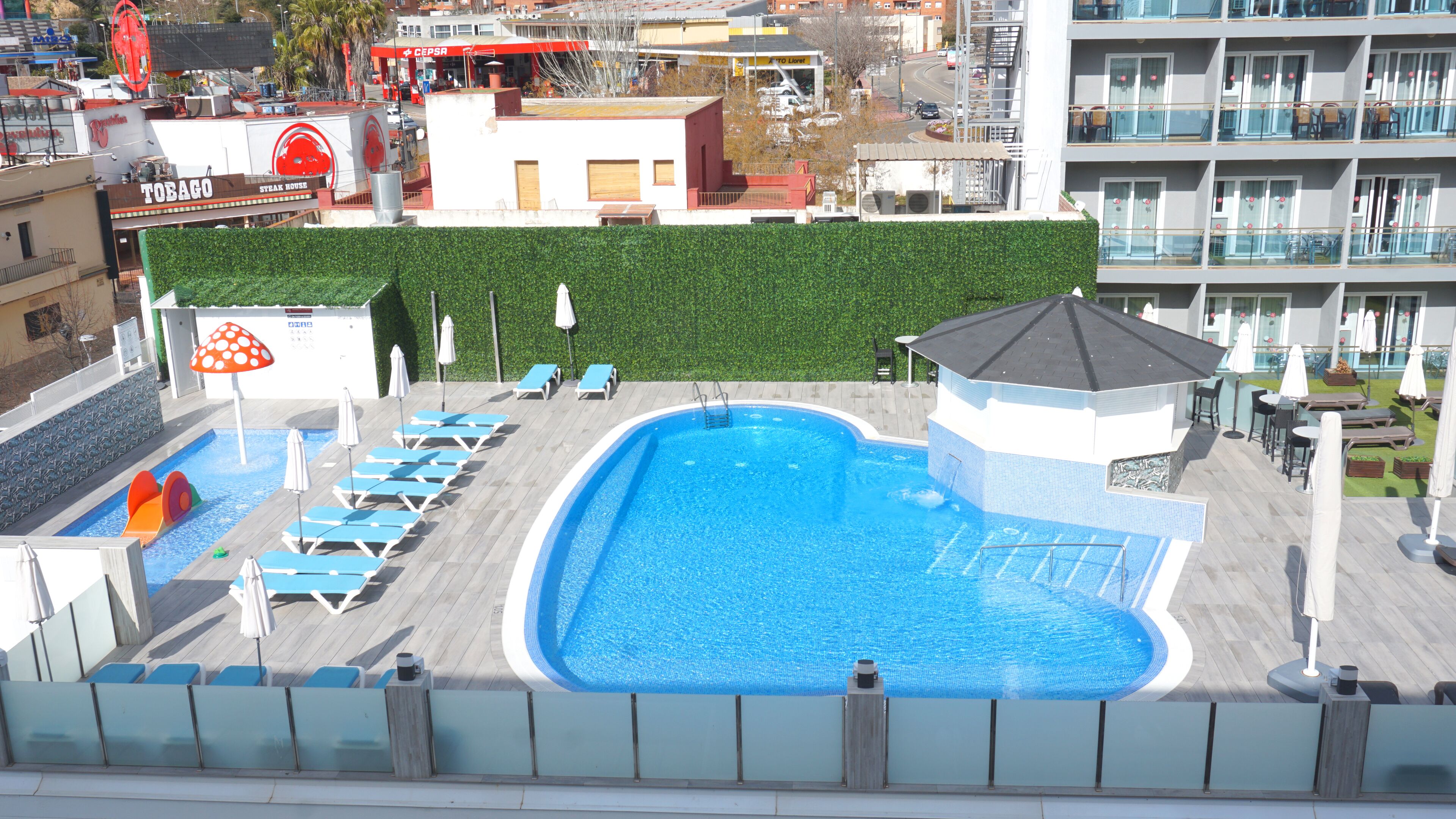 Une piscine entourée d’une haie verdoyante, avec des chaises longues et des parasols, et un bâtiment sur lequel on peut lire l’inscription « TOBAGO STEAK HOUSE ».