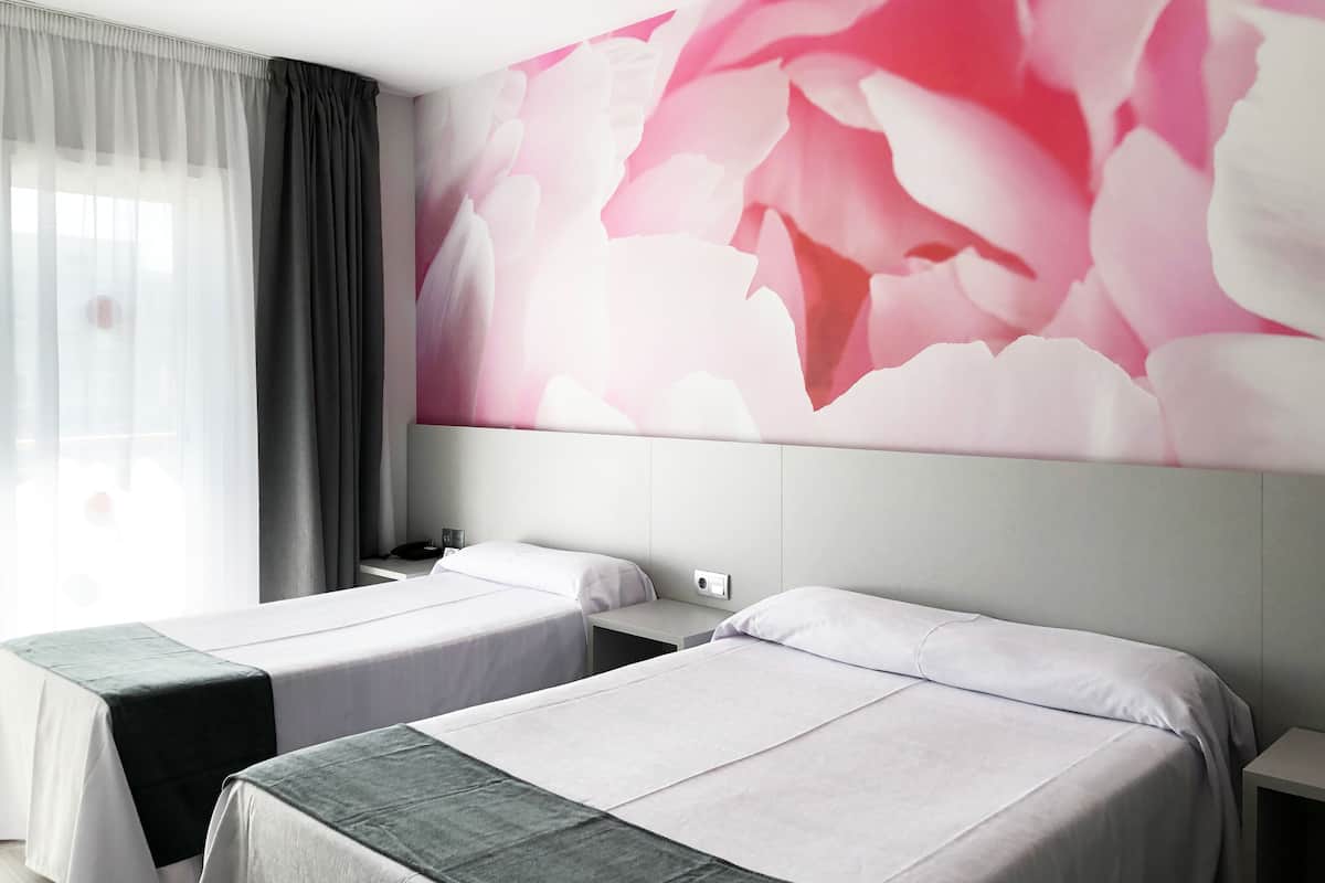 Ein Hotelzimmer mit zwei Betten, einer großen Blumenwand, einem Fenster mit durchsichtigen Vorhängen und einem Nachttisch mit Telefon.