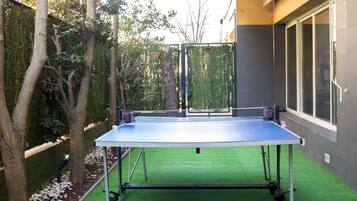 Une table de ping-pong installée dans un espace extérieur recouvert de gazon synthétique, entourée d’arbres et d’une clôture.