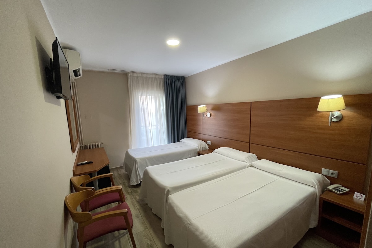 Ein Hotelzimmer mit zwei Betten, einem Schreibtisch, einem Stuhl, einem an der Wand montierten Fernseher und einem Fenster mit Vorhängen.