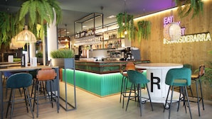 Una zona de bar con un letrero de neón que dice "EAT & DRINK DEBARRA", una barra con una máquina de café y sillas tapizadas en verde.