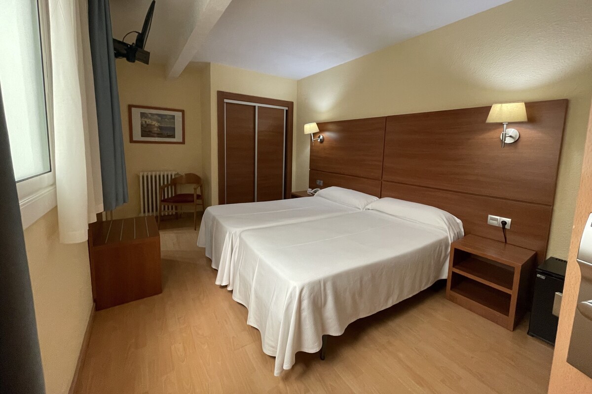 Une chambre d’hôtel avec deux lits, une tête de lit en bois, des lampes de chevet, un radiateur, une chaise et une petite table.