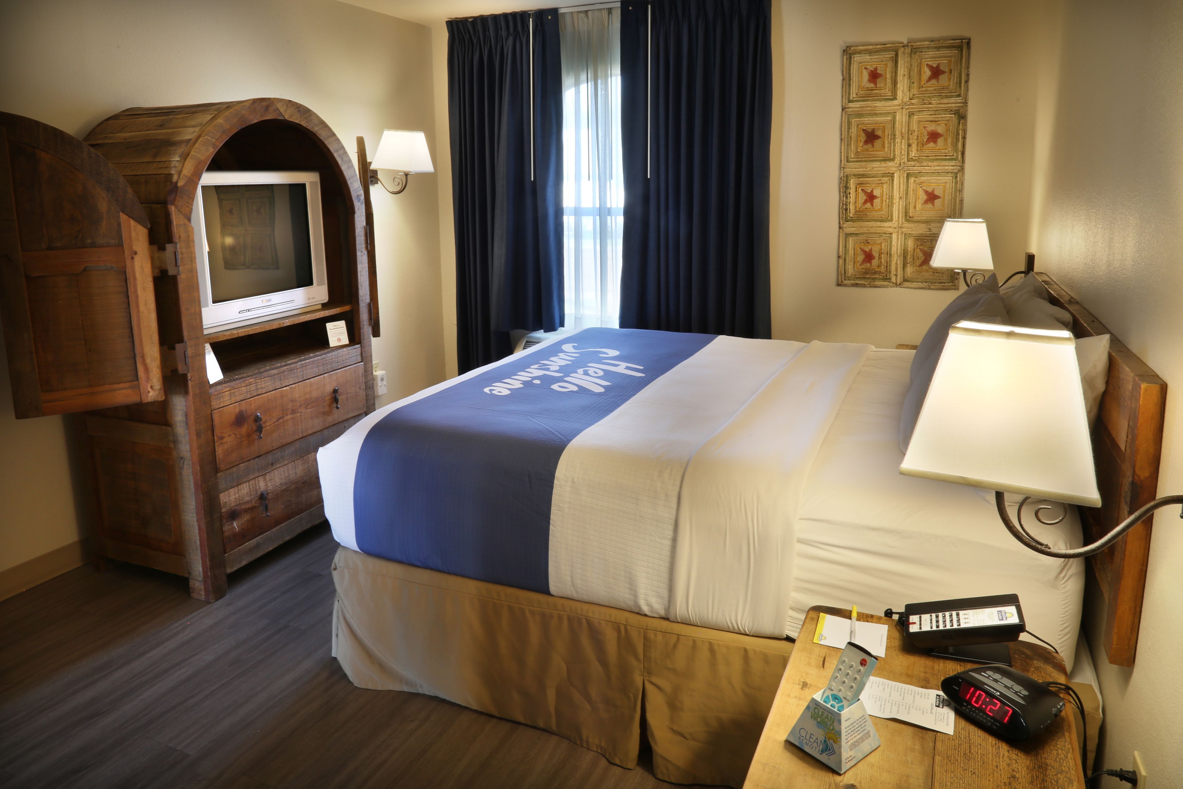 A hotel room with a bed, a wooden TV stand, a desk with a lamp, and a wall clock.