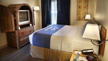 A hotel room with a bed, a wooden TV stand, a desk with a lamp, and a wall clock.