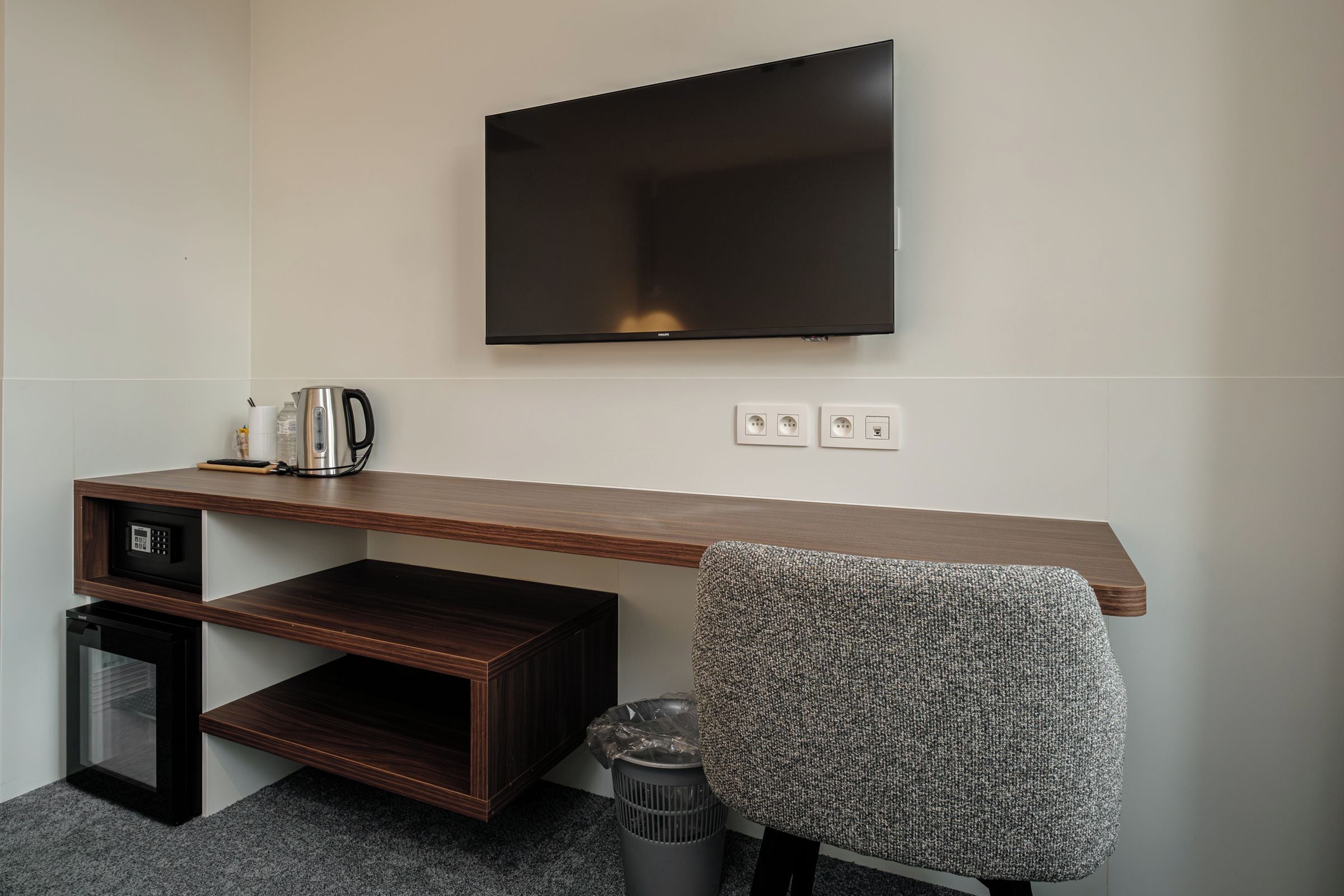 Une chambre d’hôtel moderne équipée d’un téléviseur à écran plat fixé au mur, d’un bureau en bois avec une bouilloire, d’une poubelle et d’une chaise.