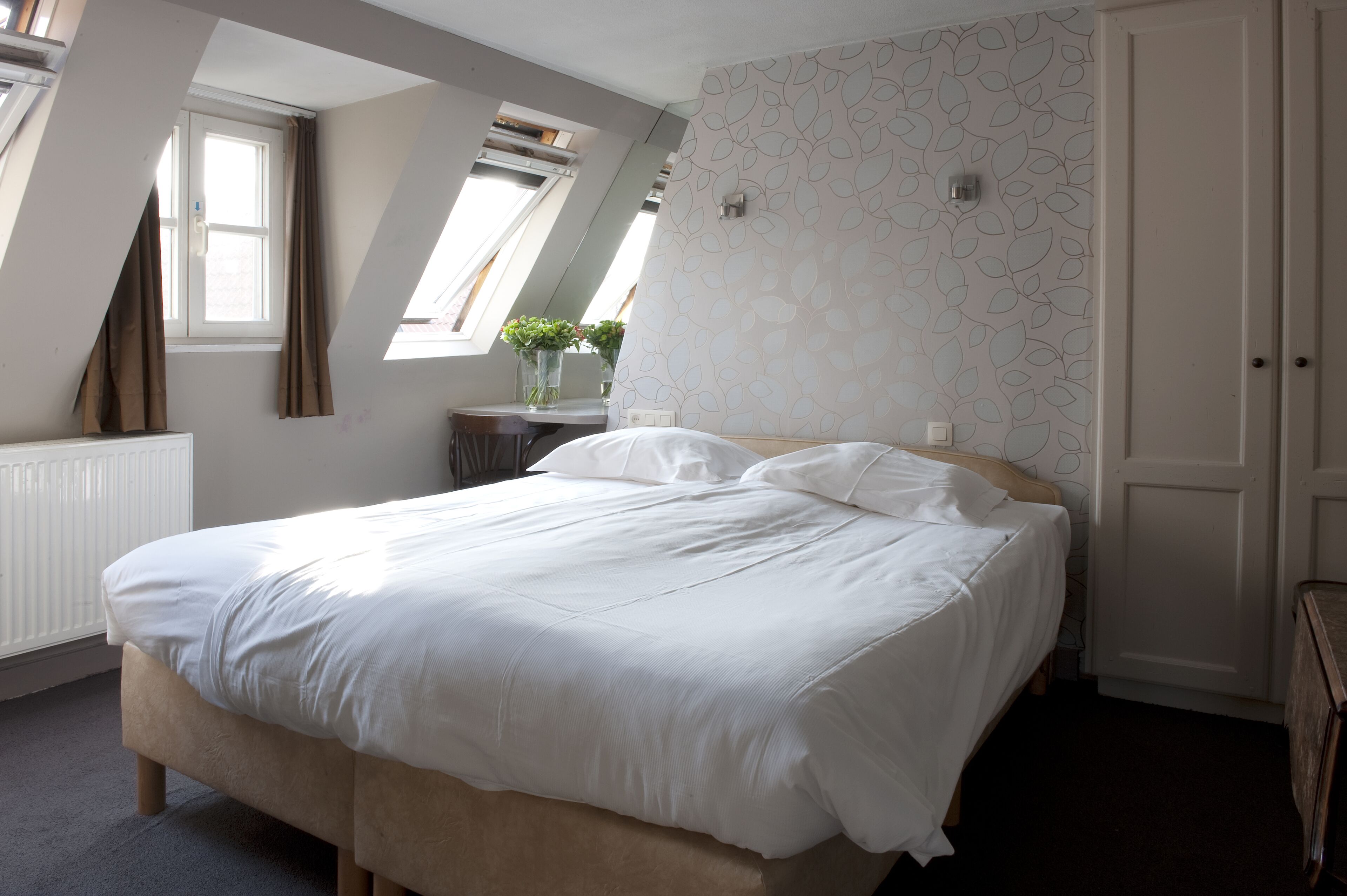 Un dormitorio con una cama grande, un radiador, una ventana con cortinas, un escritorio con una planta y una pared con un diseño decorativo.