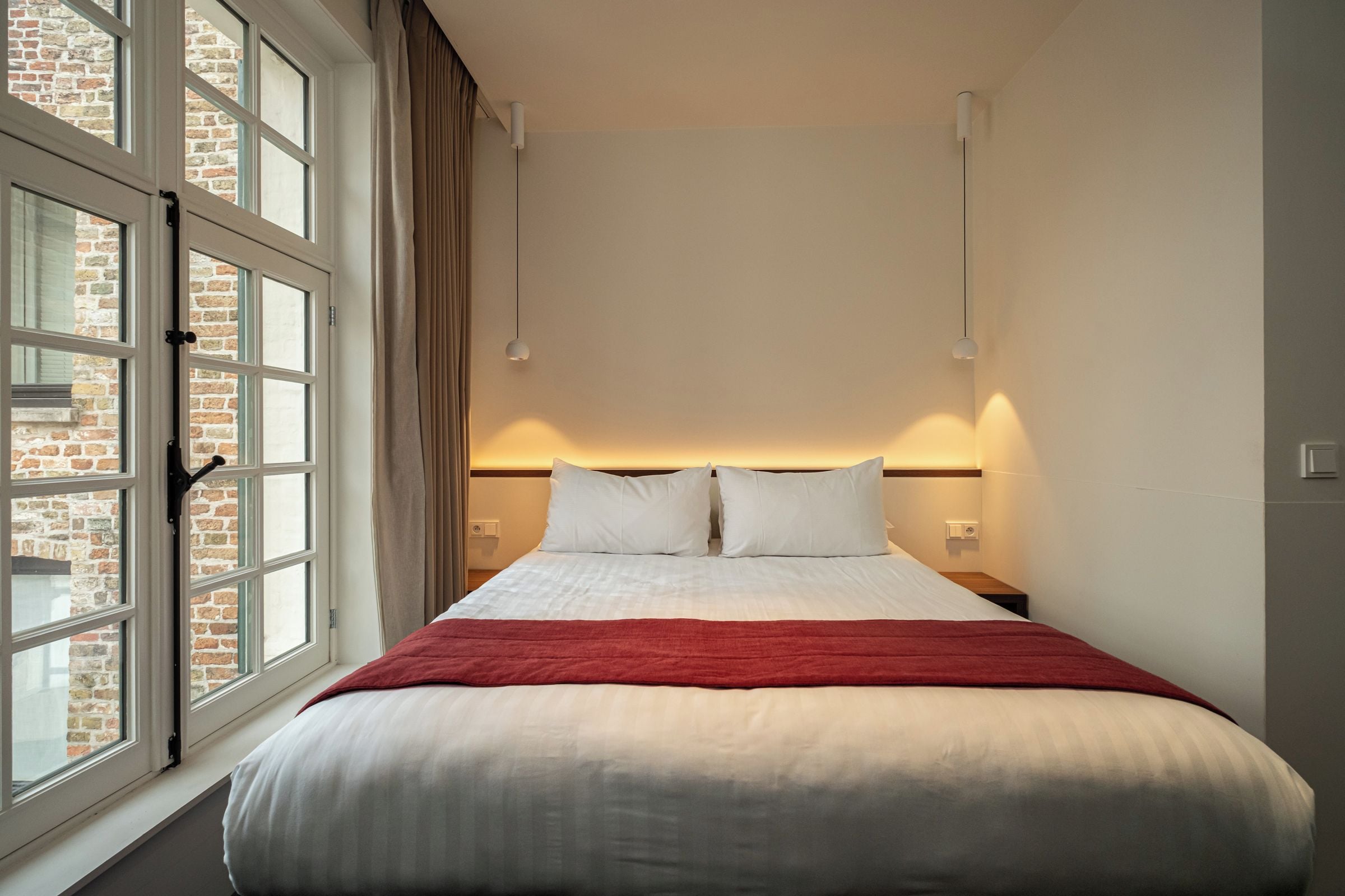 A hotel room with a large bed, a window with curtains, and a brick wall visible through the window.