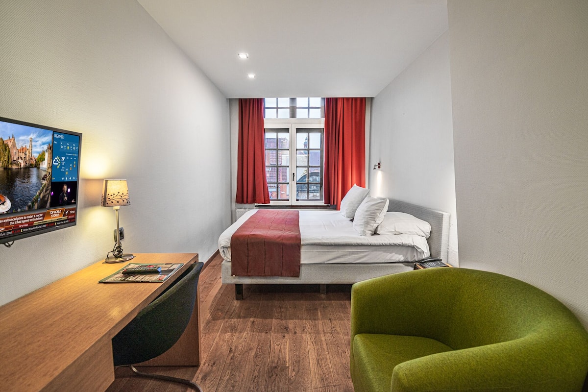 Habitación de hotel con cama, escritorio con lámpara, una silla verde y televisor mostrando un pronóstico del tiempo.