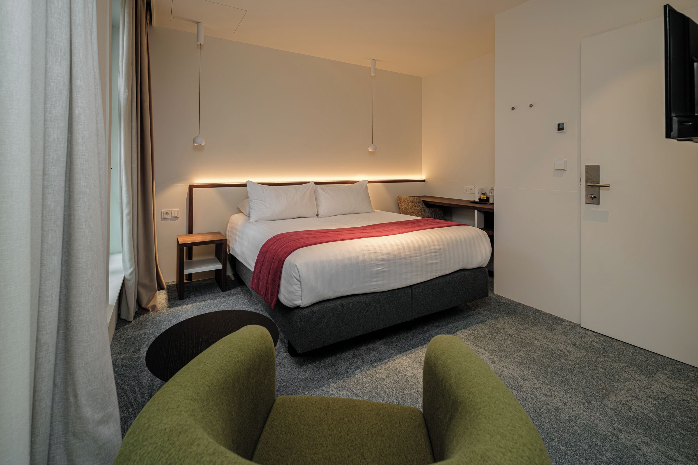 Una habitación de hotel con cama, mesita de noche, lámpara y un sillón verde.