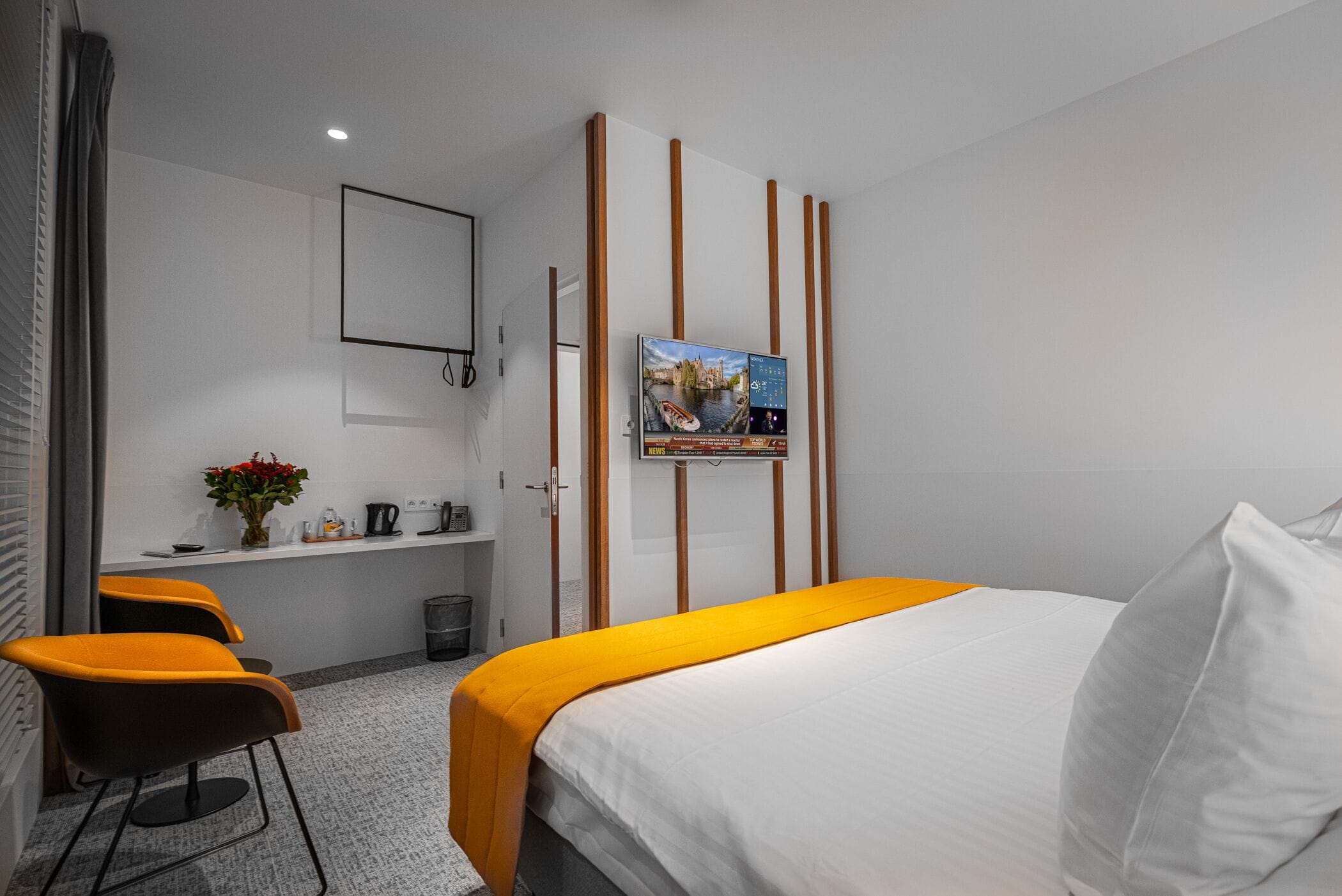 A hotel room with a bed, a desk, two chairs, a small table with a vase of flowers, and a poster on the wall.