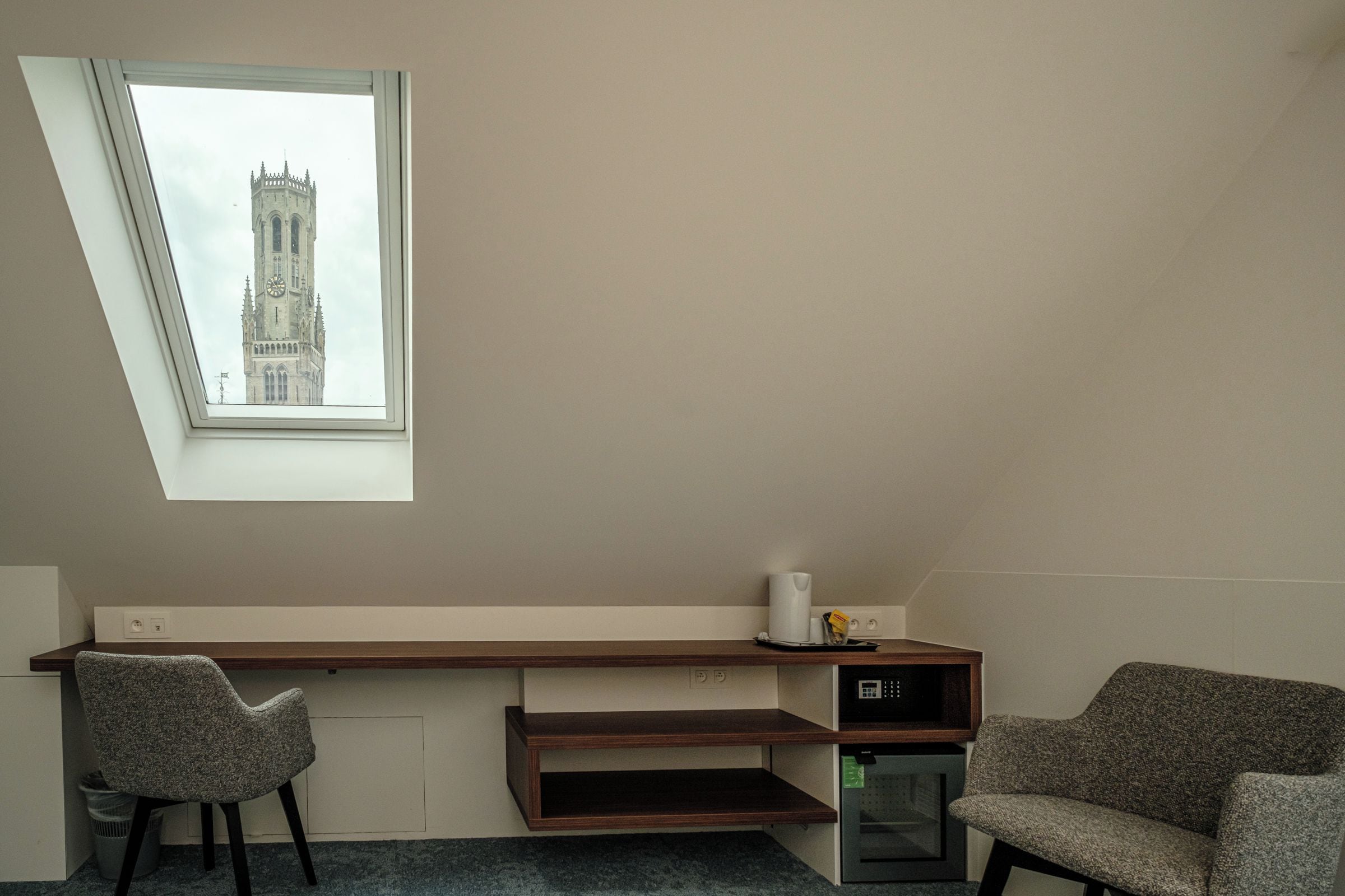 A room with a sloped ceiling, a built-in desk, two chairs, a small heater, and a view of a tower through a skylight.
