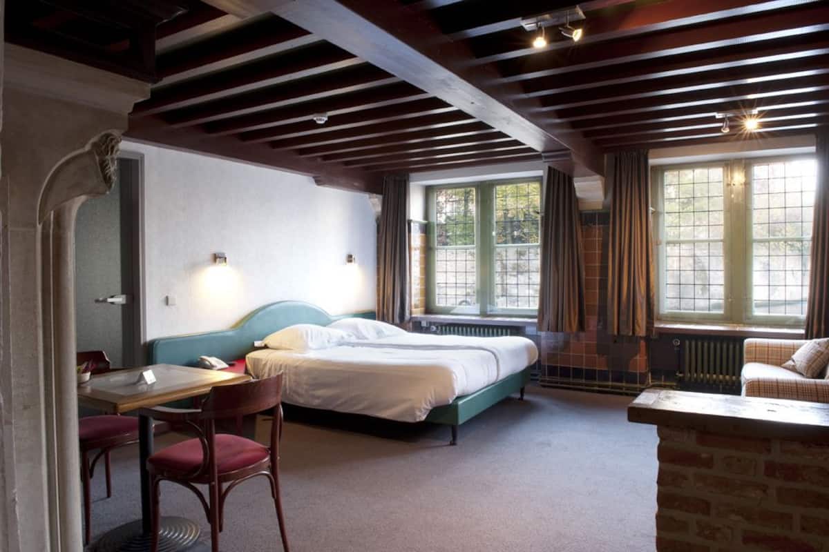 Una habitación con una cama, un escritorio y una silla. Hay ventanas con cortinas, un radiador y una pared de ladrillo.