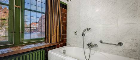Une salle de bain avec une baignoire, une fenêtre donnant sur des immeubles et un radiateur.