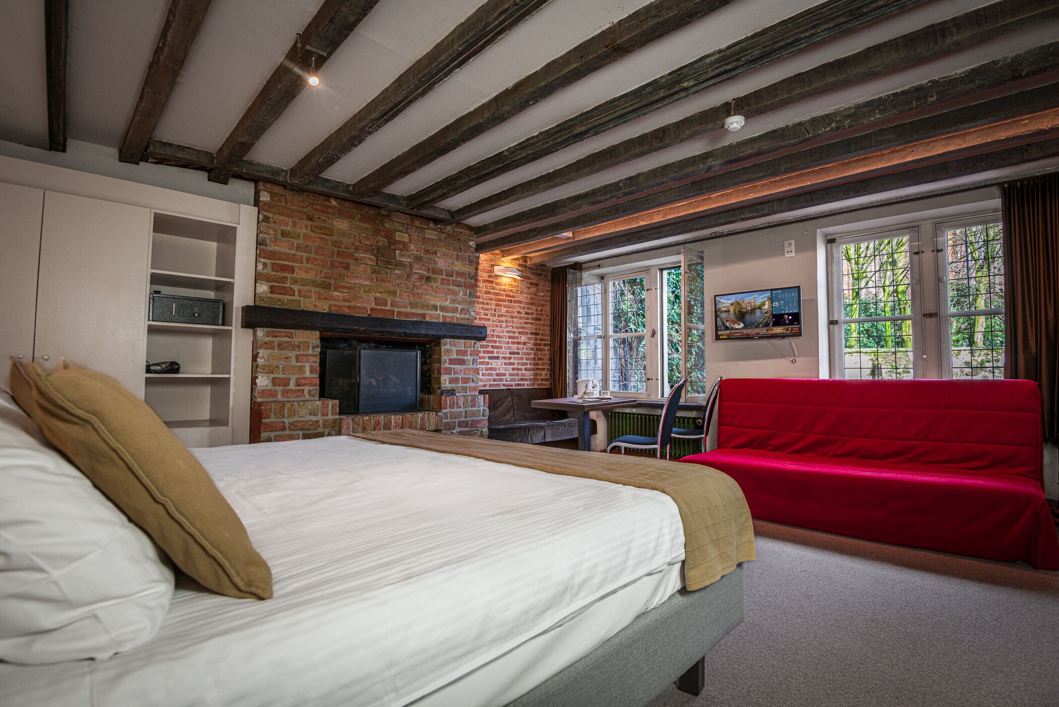 A room with a bed, a red sofa, a brick fireplace, and a TV on the wall.