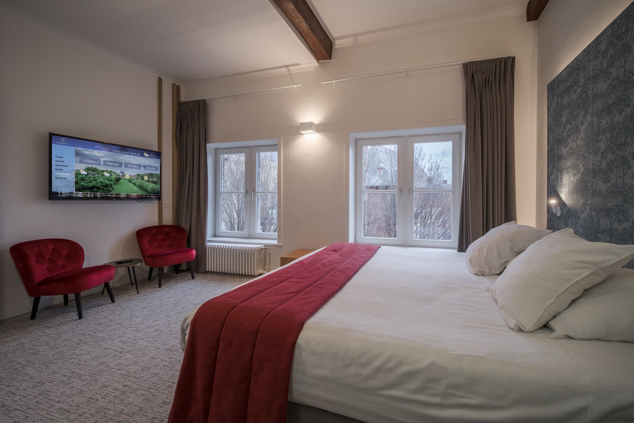 Habitación de hotel con una cama grande, dos sillones rojos, un televisor en la pared y ventanas amplias.