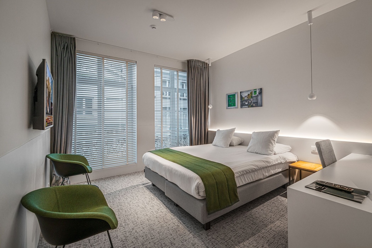 A modern hotel room with a large bed, a desk, a chair, and a window with blinds.