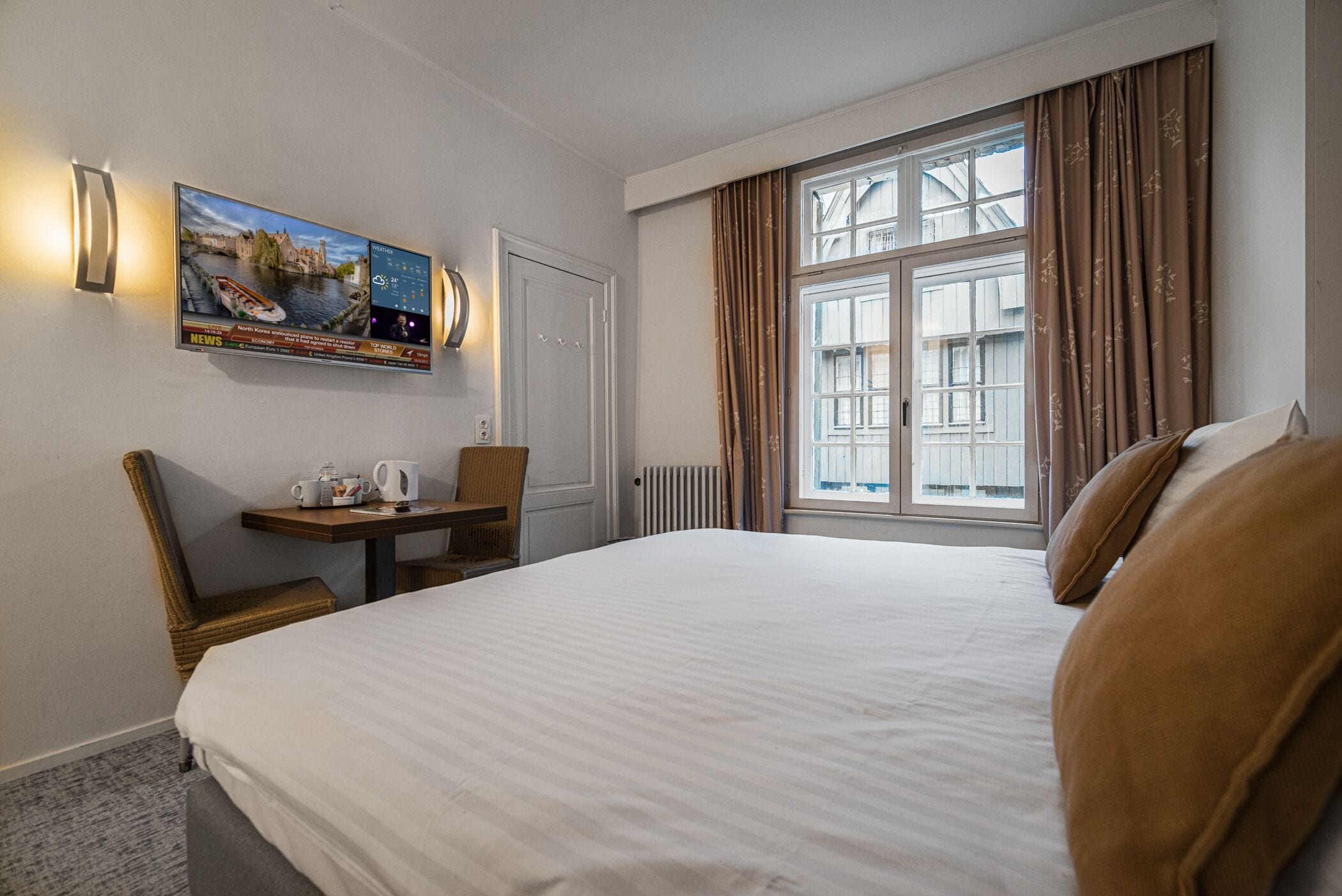 Et hotelværelse med en stor seng, et skrivebord med stol, et fjernsyn der viser nyheder og et vindue med gardiner.