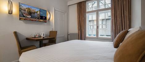 Et hotelværelse med en stor seng, et skrivebord med stol, et fjernsyn der viser nyheder og et vindue med gardiner.