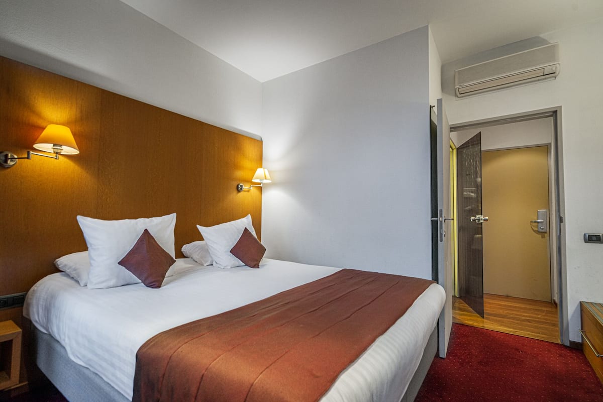 Una habitación de hotel con una cama grande, dos lámparas fijadas a la pared, un cabecero de madera, una alfombra roja y una puerta que da a un pasillo.
