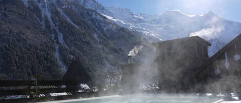 A hot spring pool with a mountainous backdrop, steam rising from the water, and a building emitting steam.