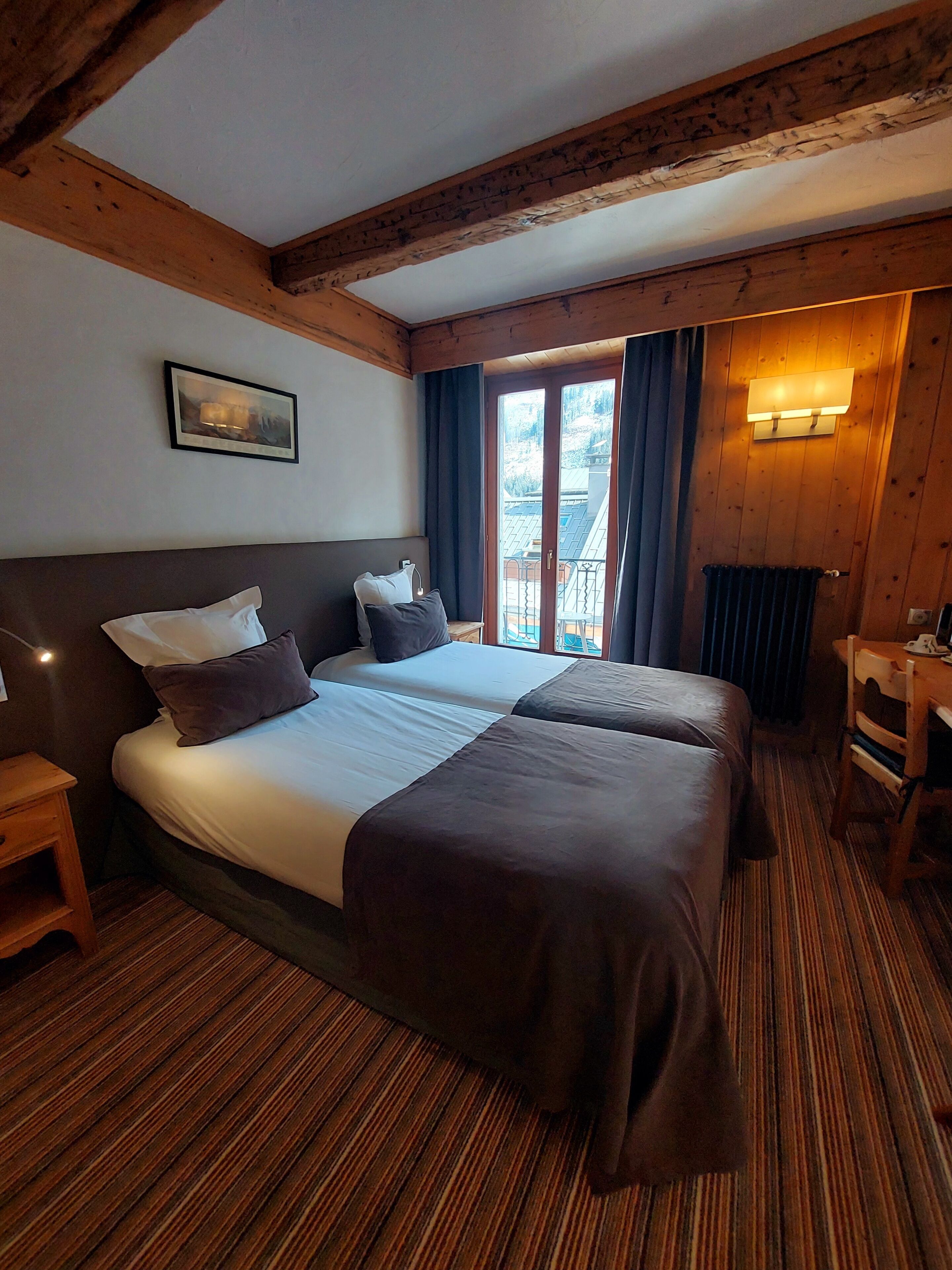 A double bed with two pillows, a bedside table, a wooden headboard, a window with curtains, and a wall-mounted light fixture.