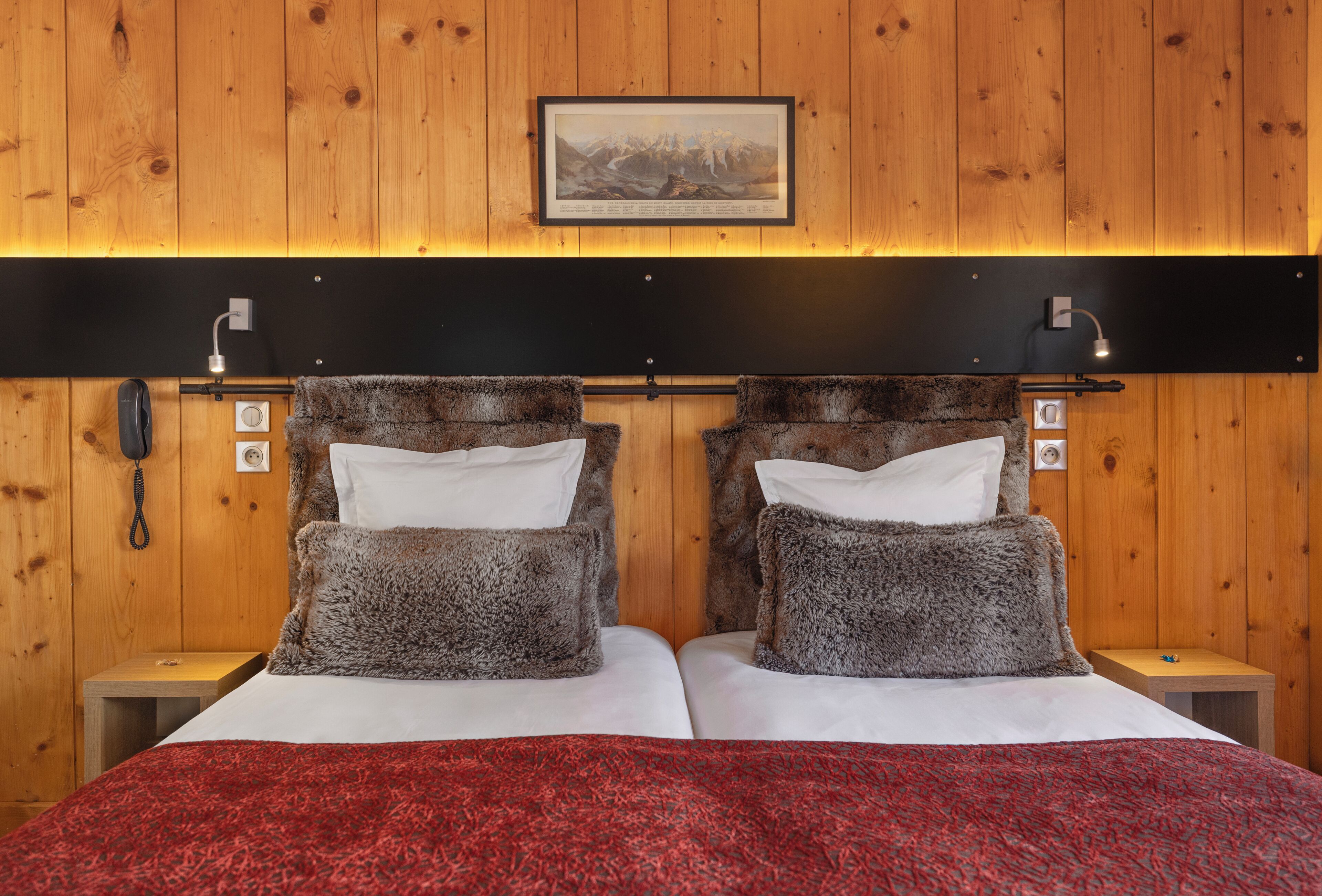 Un lit double avec une couverture rouge, deux tables de chevet avec des lampes, un tableau encadré au mur et un lambris en bois.
