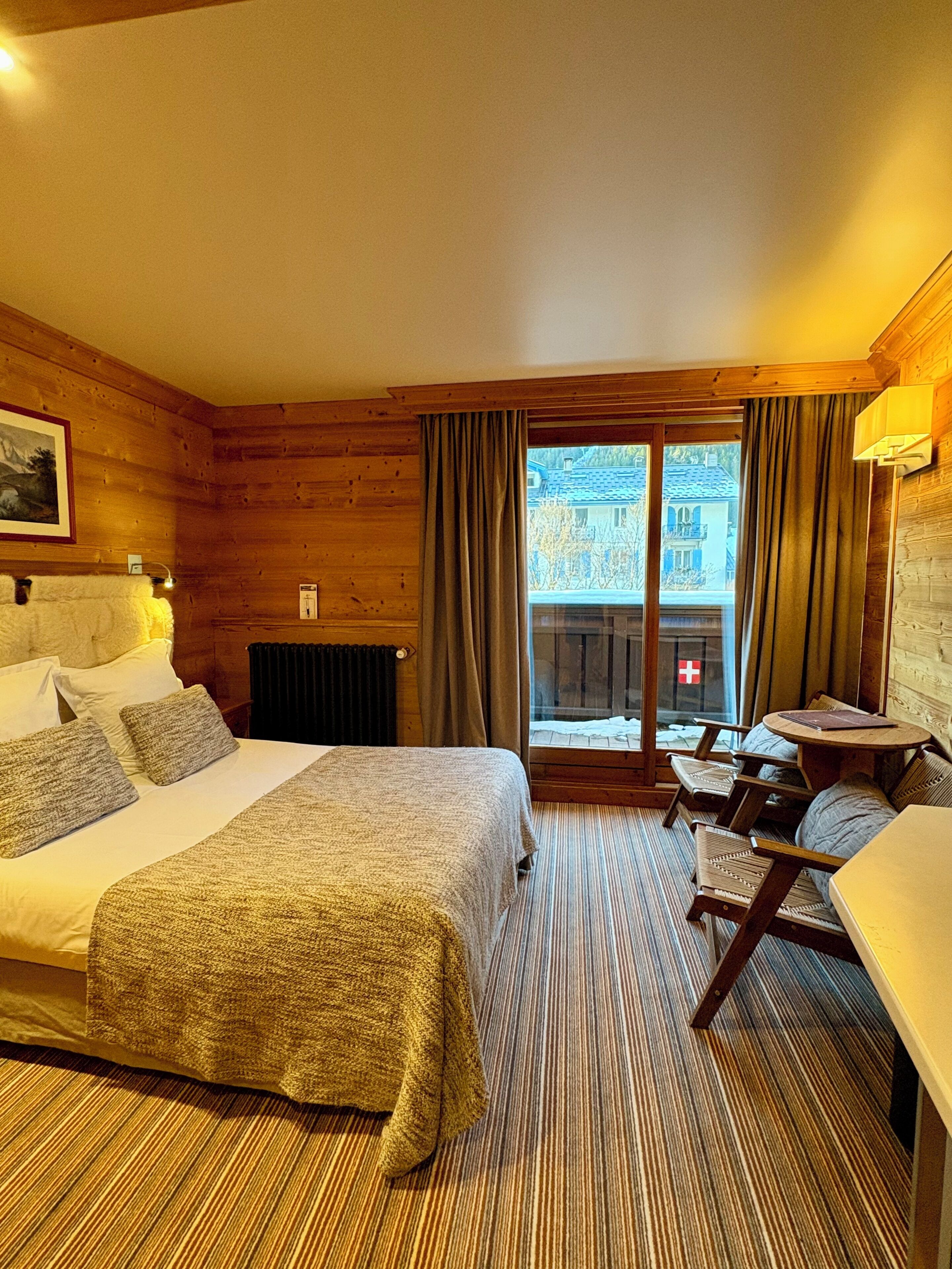 A hotel room with a bed, a small table, and a view of the outside.
