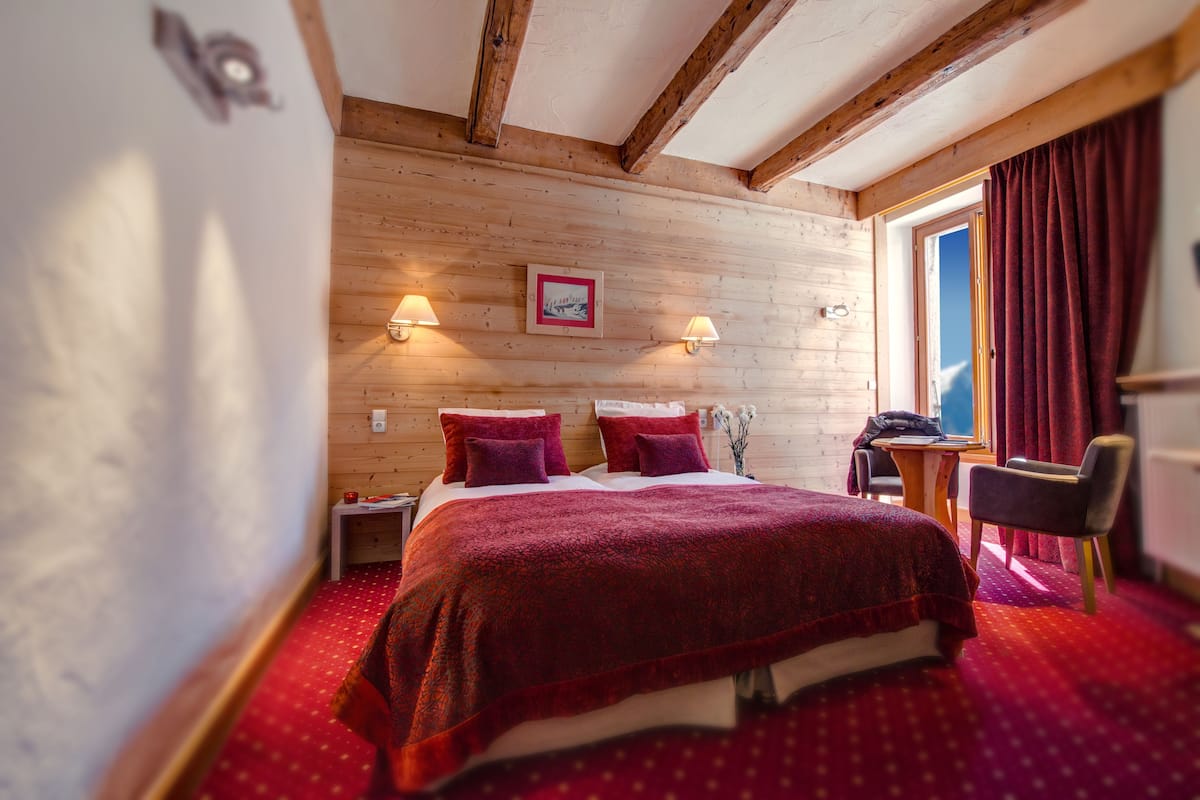 Ett sovrum med ett träpanelat tak, en stor säng med röda sängkläder, två sänglampor, ett litet bord med en stol och ett fönster med röda gardiner.