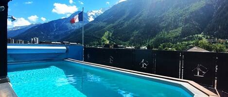 Una piscina con agua azul cristalina, rodeada por una cubierta de madera y una valla metálica. Al fondo, montañas y un asta con una bandera.