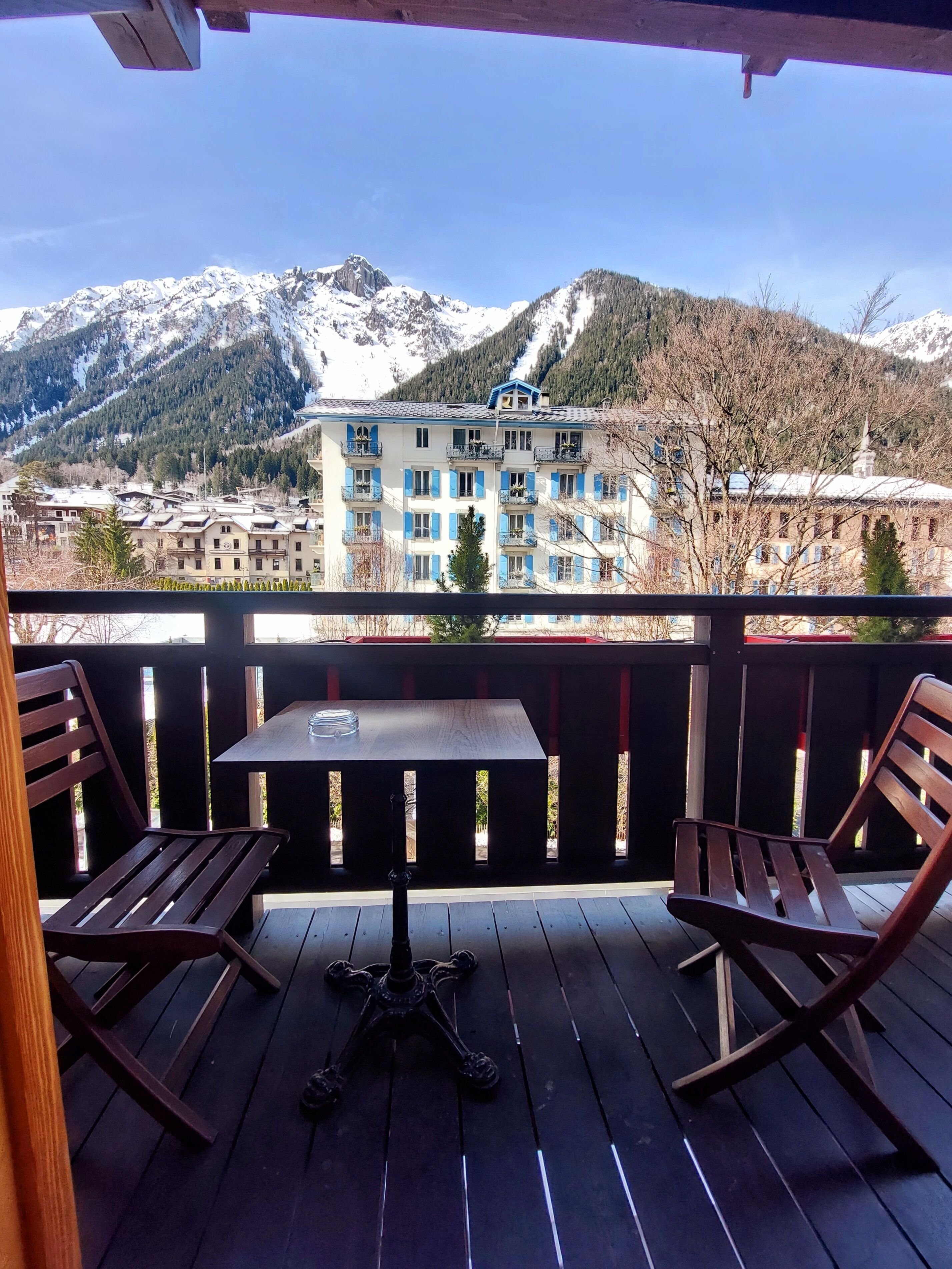 Un balcón con dos sillas de madera y una mesa, con vistas a un paisaje montañoso y un edificio con contraventanas azules.