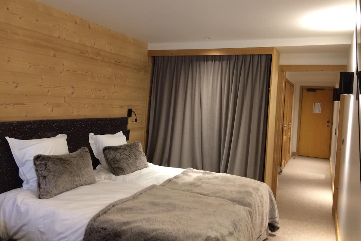 Ein Hotelzimmer mit einem hölzernen Kopfteil, einem Bett mit weißer Bettwäsche und zwei grauen Kissen, einem grauen Vorhang und einer Holztür.
