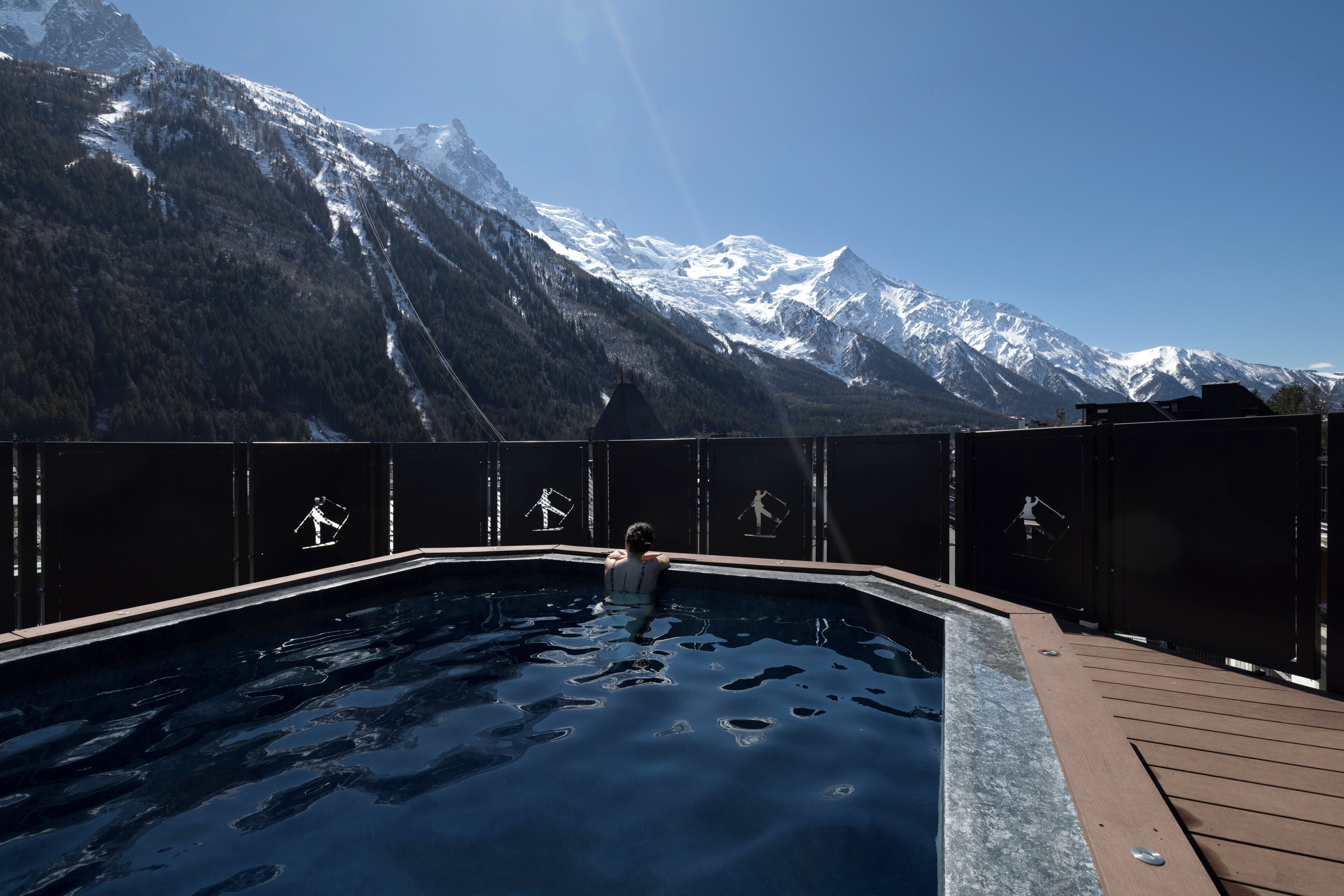 En swimmingpool med ett trädäck, omgiven av ett staket av metall, och en bergig bakgrund.