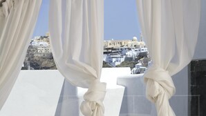 A room with a large window, white curtains tied back, and a bed with a beige hat, glasses, and a camera on it. - Blue Angel Villa (Santorini)