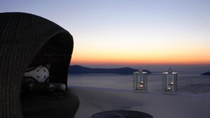 A wicker lounge chair with a patterned cushion, a small round table, and two lit lanterns on a balcony overlooking a beach at sunset. - Blue Angel Villa (Santorini)