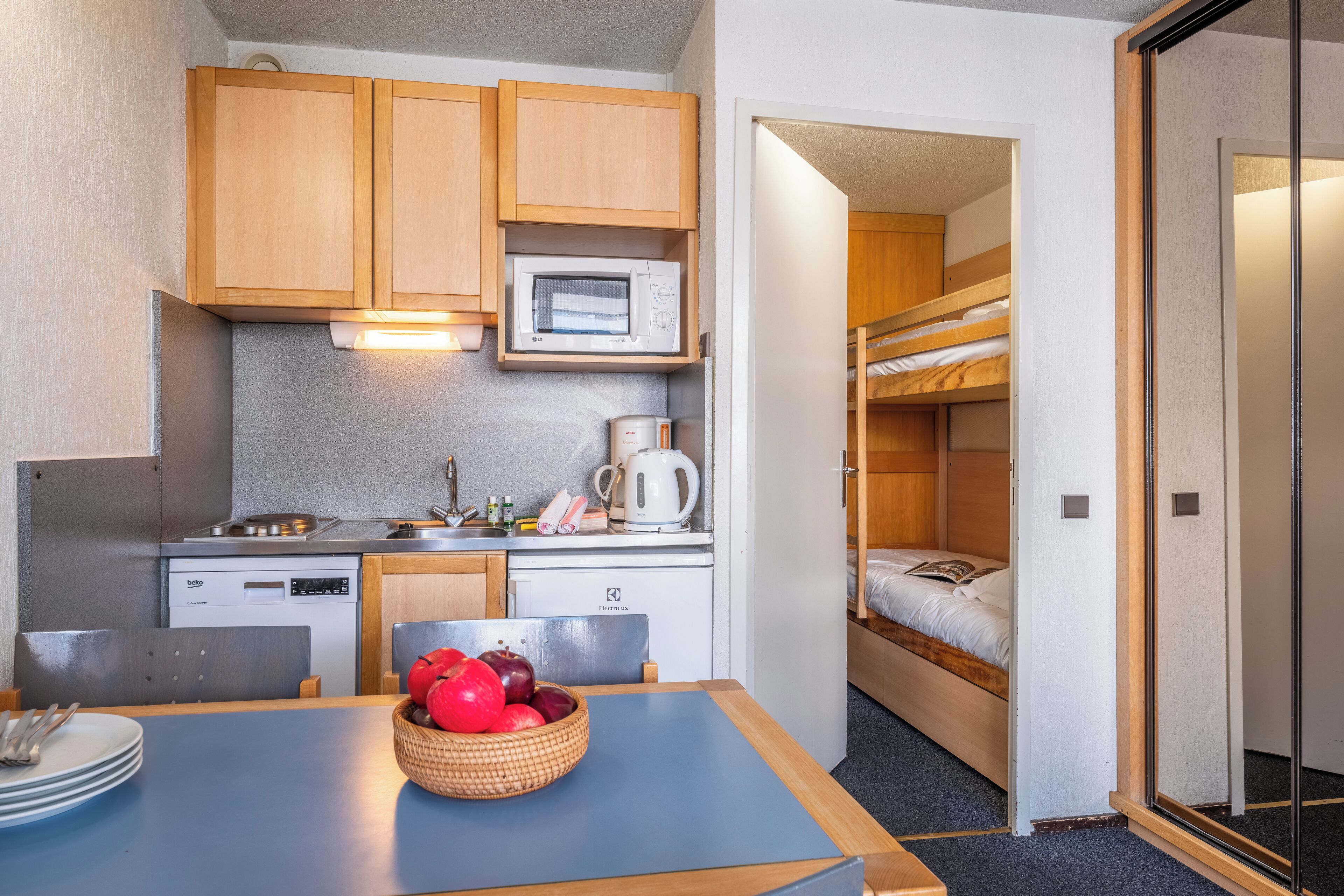 A compact kitchen with a microwave, coffee maker, and a sink. There is a dining table with a bowl of fruit and a chair. A bunk bed is visible in the background.