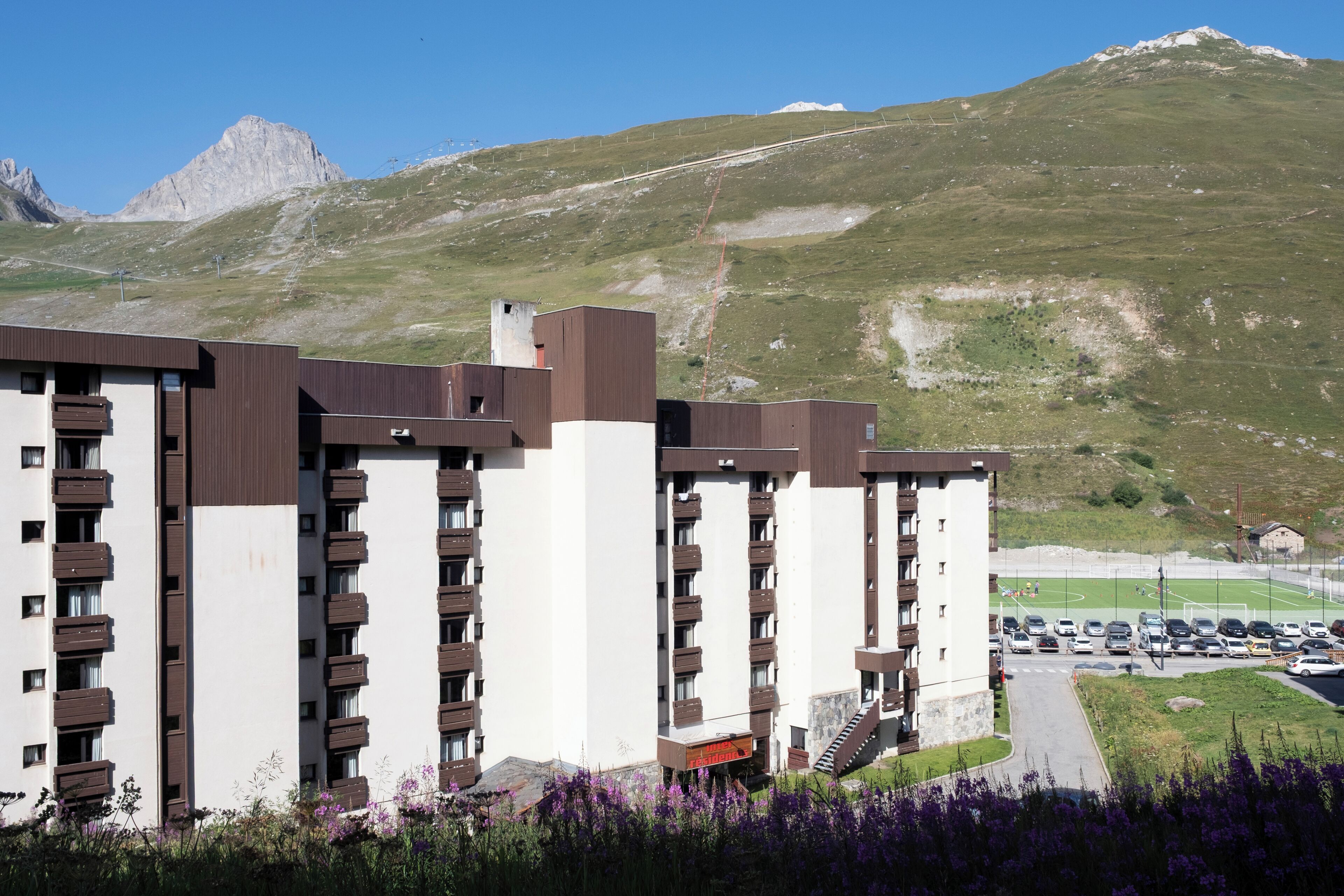 Een meerlaags hotel met balkons, een parkeerplaats en een bergachtige achtergrond.