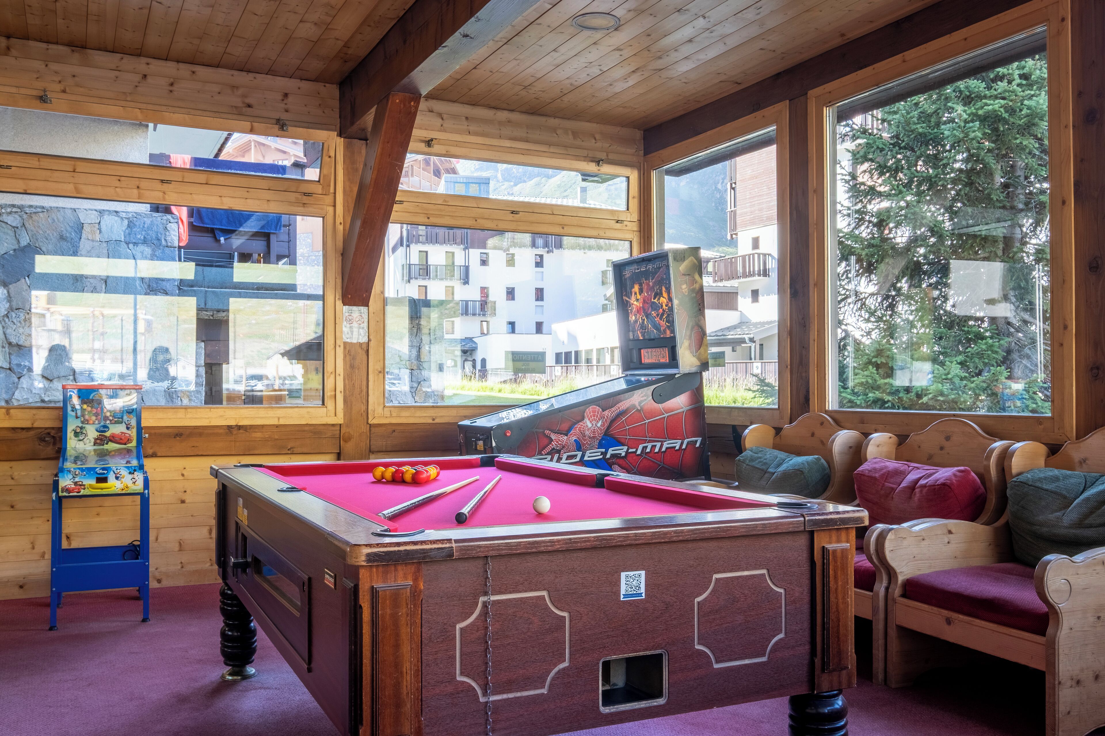 A pool table with a Spider-Man theme, wooden furniture, and large windows with a view of buildings and trees.