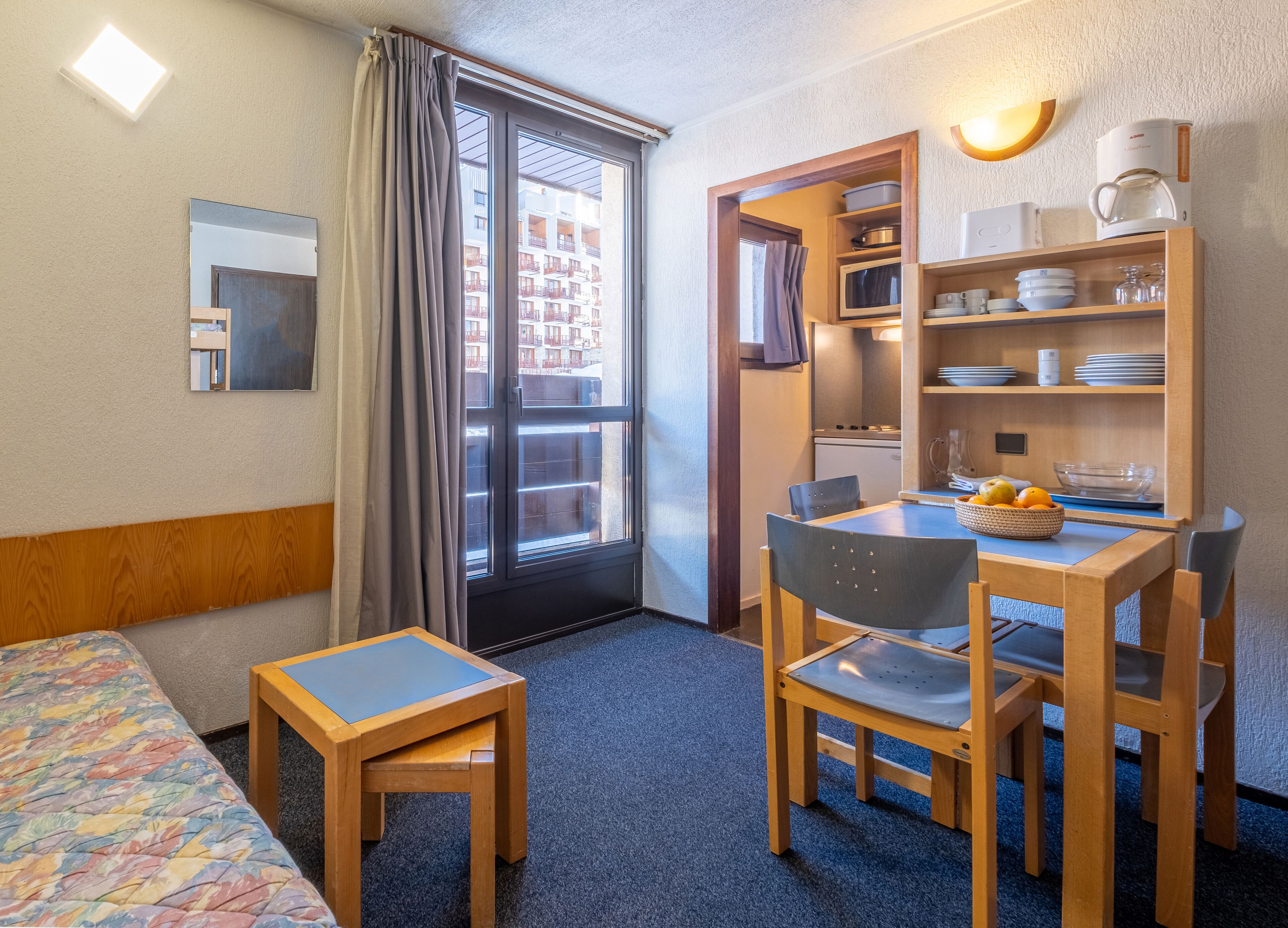 A hotel room with a bed, a small table, a chair, a dining table with chairs, a microwave, a coffee maker, and a view of the outside balcony.