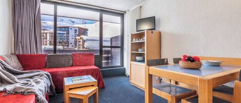 Hotelkamer met een rode bank, een klein houten tafeltje, een tv op een houten standaard en uitzicht op een besneeuwde berg.